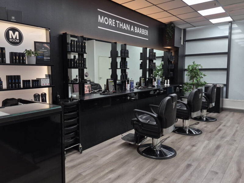 Parturiliikkeen sisäkuva: kolme mustaa parturituolia pitkän mustan työpöydän ja peilien edessä, hyllyt hiustuotteineen, viherkasvi ja kyltti "MORE THAN A BARBER" peilien yläpuolella, vaalea puulattia ja ruutukatto.