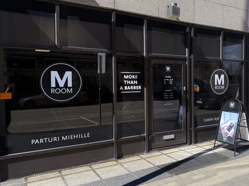 Torikadun näköala Joensuussa: M Room -miesten parturin musta lasijulkisivu, lasiovet ja ikkunat joissa näkyvät logot "M ROOM" ja teksti "MORE THAN A BARBER" sekä kadulla oleva mainoskyltti.