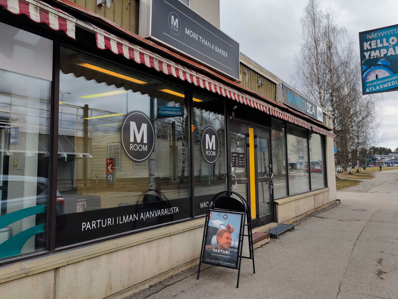 Kauppakadun kivijalkaliikkeen julkisivu: M Room -parturiliike ikkunalogoilla ja punavalkoisella markiisilla, oven edessä mainoskyltti hiusleikkauskuvalla, tyhjä jalkakäytävä ja taustalla lehtipuita ja taloja.