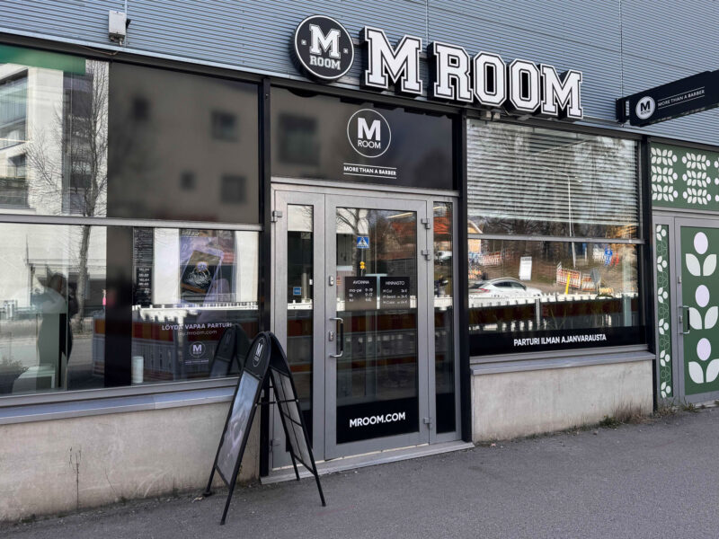 M ROOM -liikkeen lasijulkisivu Kauniaisissa: suuri M ROOM -kyltti oven yläpuolella, lasiovet ja näyteikkunat joissa heijastuksia, jalkakäytävällä A-telinemainos.