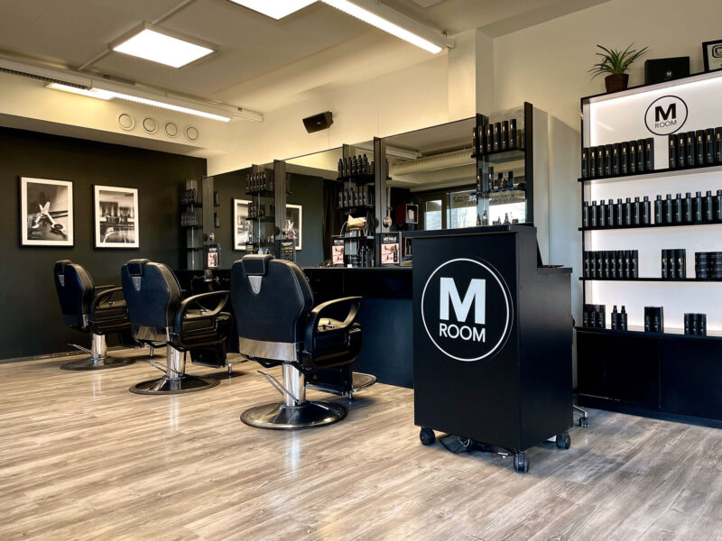 Modernin M Room -parturiliikkeen sisätila: kolmen mustan parturituolin rivi peilin edessä, hyllyt täynnä parturituotteita, etualalla musta kassatiski, jossa valkoinen M ROOM -logo, vaaleaparketti, seinällä mustavalkoisia kuvia ja katon valaisimet.