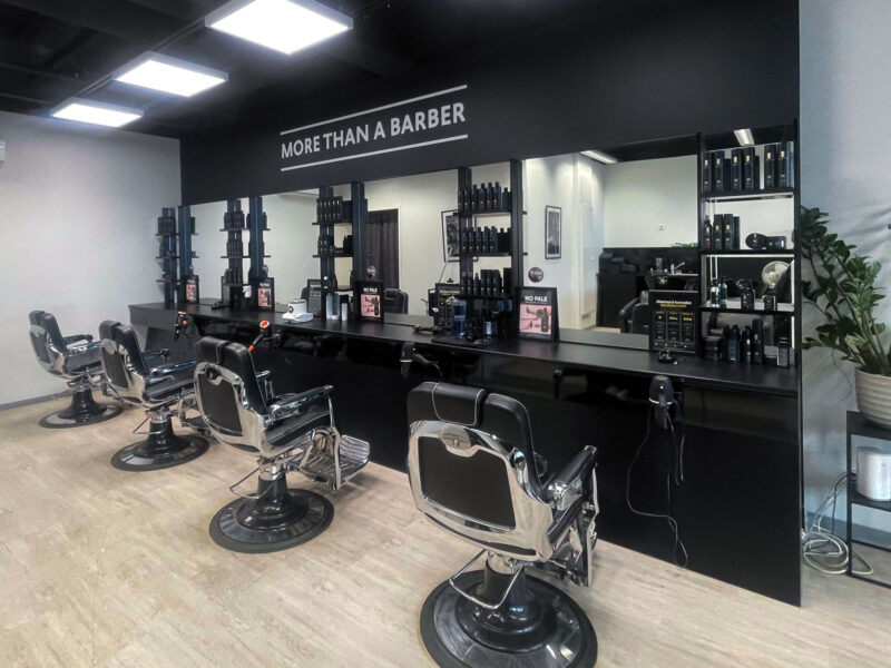 Sisäkuva modernista parturiliikkeestä, jossa rivissä mustia parturituoleja peilien edessä, pitkä musta työtaso leikkausvälineineen ja tuotteineen, yläpuolella teksti MORE THAN A BARBER, vaalea lattia ja kirkkaat katto‑valaisimet.