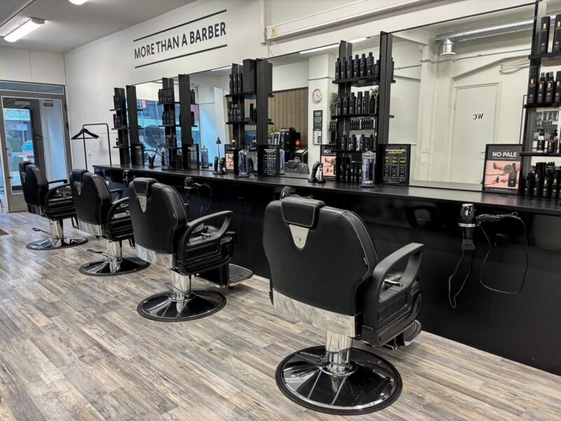 Nykyaikainen parturiliike, rivissä mustat parturituolit peilien edessä, pitkä musta työtaso ja hyllyt täynnä hiustuotteita, vaalea laminaattilattia ja seinässä teksti "MORE THAN A BARBER".