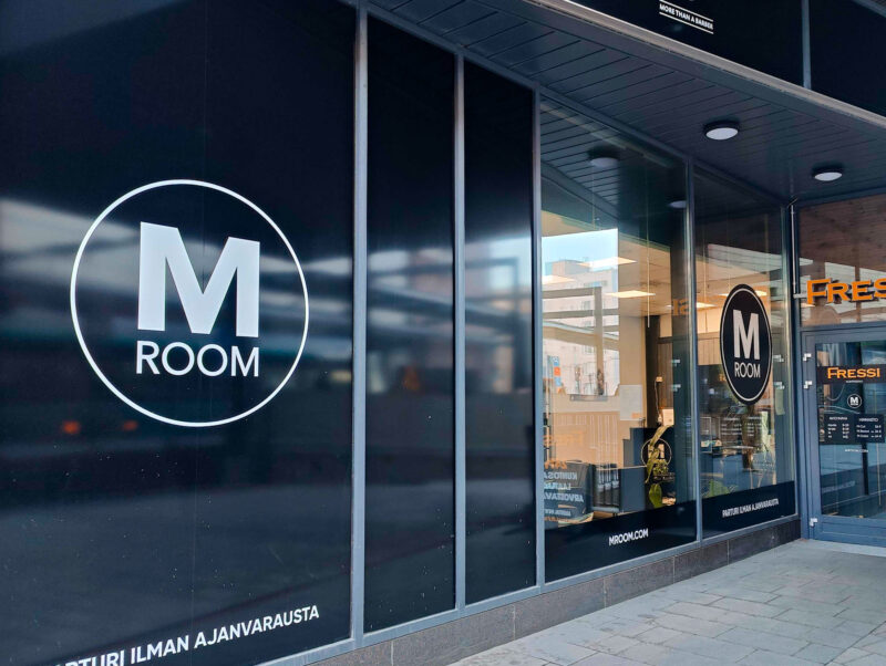 Parturiliikkeen julkisivu ja sisäänkäynti Espoon Panorama Towerissa: mustiin puitteisiin ja suurille ikkunoille sijoitetut valkoiset ympyrälogot "M ROOM", näkyvissä valaistu myymälän sisätila, ikkunan heijastuksia ja jalkakäytävä.