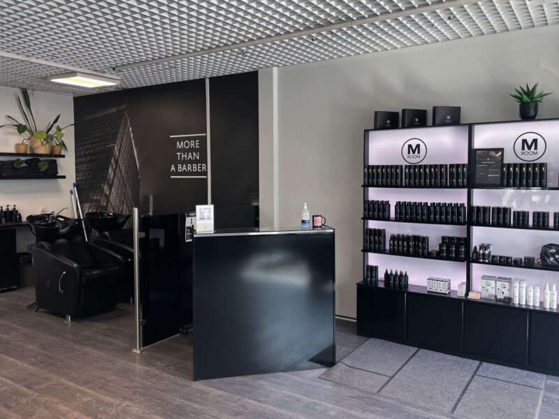 Sisäkuva Porvoon M Room -parturiliikkeestä: musta vastaanottotiski etualalla, vasemmalla pesuistuimet sekä suuri seinäkuva, jossa teksti "MORE THAN A BARBER", oikealla valaistut hyllyt täynnä hiustuotteita ja alaosassa kaapit, katossa ruudukkomallinen kattopaneeli ja näkyvissä muutama viherkasvi.