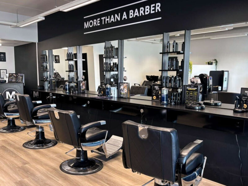 Modernin parturiliikkeen sisätila: kolme mustaa parturituolia peilien edessä, peilien yläpuolella teksti MORE THAN A BARBER, hyllyt täynnä hiustuotteita, vaalea puulattia