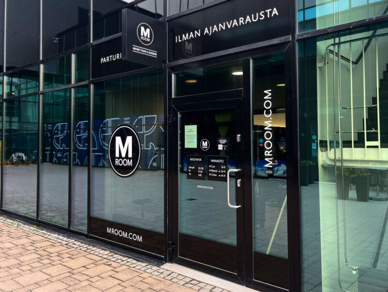 Lasinen sisäänkäynti parturiliikkeeseen M Room Tampereen Lielahdessa; oven ja ikkunoiden näkyvillä M Room -logo, tekstit "PARTURI", "ILMAN AJANVARAUSTA" ja "MROOM.COM" sekä heijastuksia kadusta.