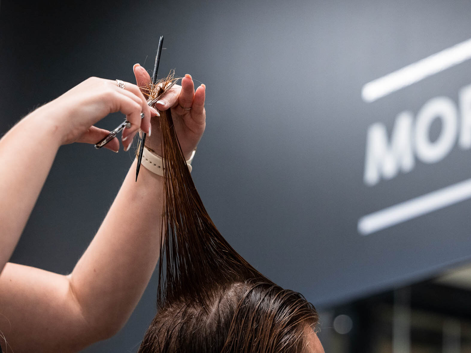 Lähikuva parturin käsistä leikkaamassa asiakkaan hiuksia saksilla, yksi hiussäie nostettuna ylös ja taustalla epäterävänä osa kylttiä, jossa lukee "MORE THAN A BARBER".