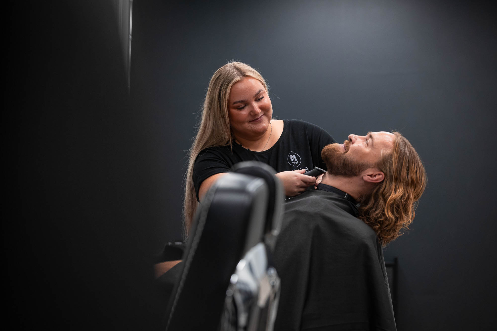 Naisparturi hymyilee rajatessaan pitkähiuksisen miehen partaa koneella, asiakas nojaa taaksepäin parturituolissa tumman taustan edessä.